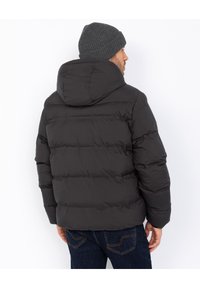 Mand iført en sort puffjakke med hætte og en mørkegrå strikket hue, stående og vendt væk fra en ensfarvet hvid baggrund.