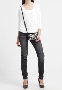 Blouse blanche à manches enveloppantes, en tissu léger ; jeans noirs, coupe slim ; sac bandoulière rayé noir et blanc assorti ; escarpins gris.