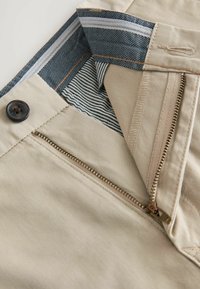 Beige nadrág félig lehúzott sárgaréz cipzárral, fekete gombbal és belső kék-fehér csíkos anyagú derékrésszel.