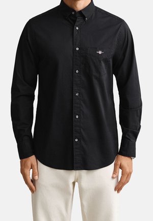 Homme portant une chemise noire à manches longues boutonnée avec une poche poitrine et un pantalon beige, debout les bras détendus.