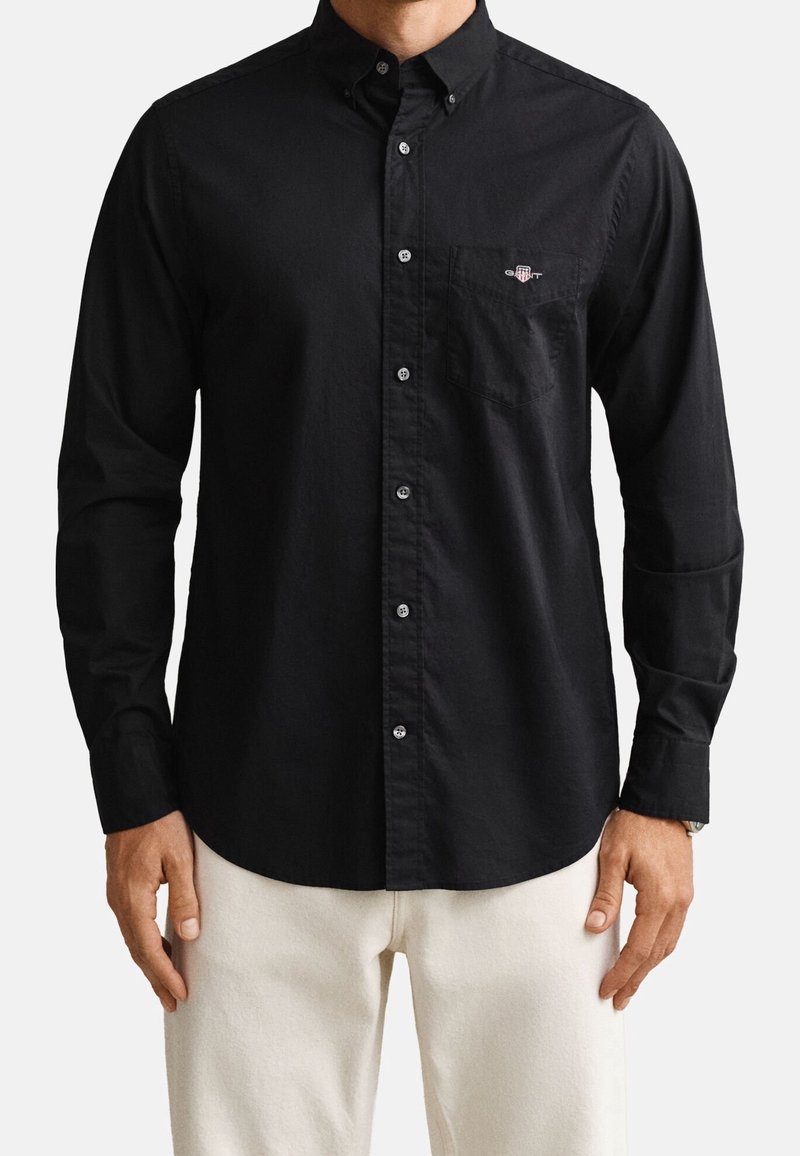 Homme portant une chemise noire à manches longues boutonnée avec une poche poitrine et un pantalon beige, debout les bras détendus.