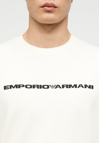 Κοντινό πλάνο ενός ατόμου που φοράει λευκό πουκάμισο με την επιγραφή "EMPORIO ARMANI" μαύρο χρώμα στο στήθος.