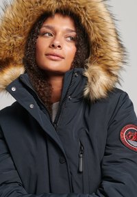 Marineblauwe parka met een nepbont capuchon, rits- en drukknoopsluiting, en een patchdetail op de mouw. Gladde textuur en geïsoleerd ontwerp.