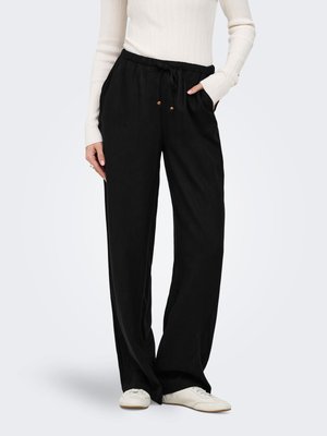 Personne portant un pantalon noir ample avec taille à cordon, un haut blanc à manches longues côtelé et des baskets blanches, debout avec une main dans la poche.