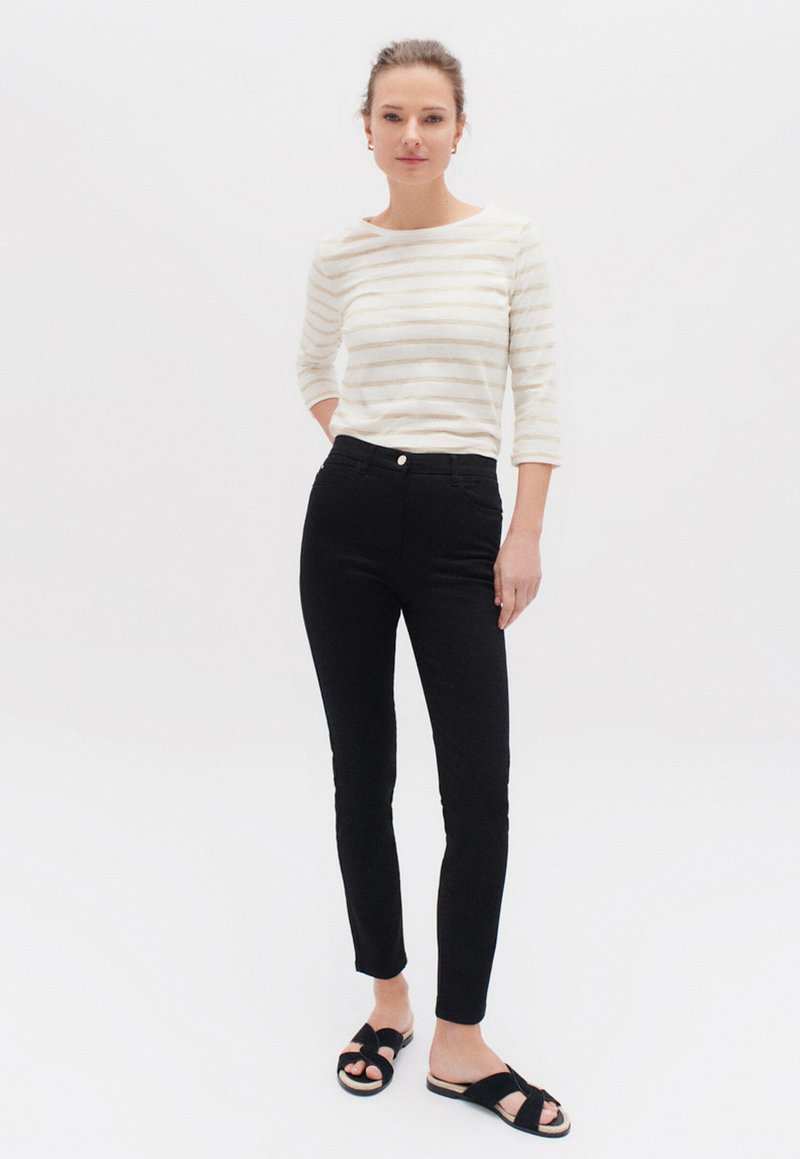 Top a maniche lunghe bianco e beige a righe con collo rotondo, abbinato a jeans skinny neri a vita alta e sandali neri a fascia.