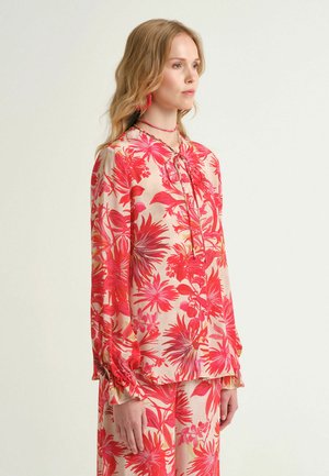 Femme portant un ensemble assorti blouse et pantalon à fleurs roses et rouges, debout de profil devant un fond clair uni.