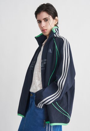 Junge Frau mit kurzen dunklen Haaren, die eine marineblaue Adidas-Jacke mit grüner Einfassung, ein weißes gemustertes Hemd und einen Jeansrock trägt und vor weißem Hintergrund posiert.