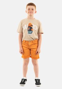 T-shirt beige con grafica di orso e testo "Fat Boy"; pantaloni corti arancioni; sneakers nere con strisce bianche; vestibilità casual e orlo arrotolato.
