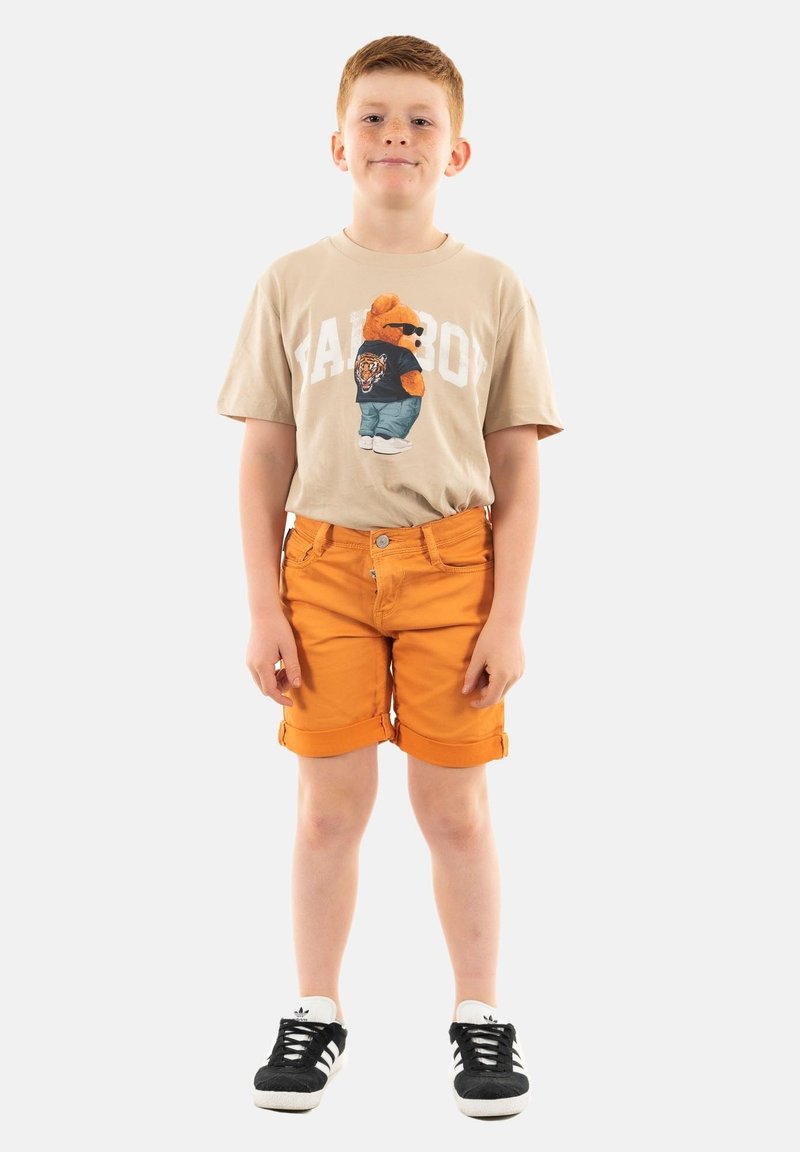 T-shirt beige con grafica di orso e testo "Fat Boy"; pantaloni corti arancioni; sneakers nere con strisce bianche; vestibilità casual e orlo arrotolato.