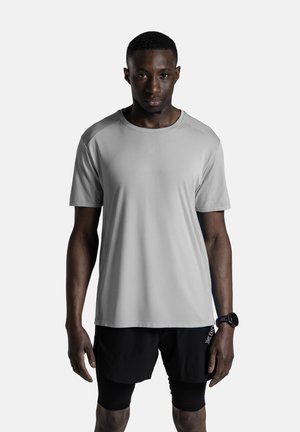 Fit man draagt lichtgrijs sportshirt, zwarte short en smartwatch, staat tegen een effen achtergrond, kijkt recht vooruit.