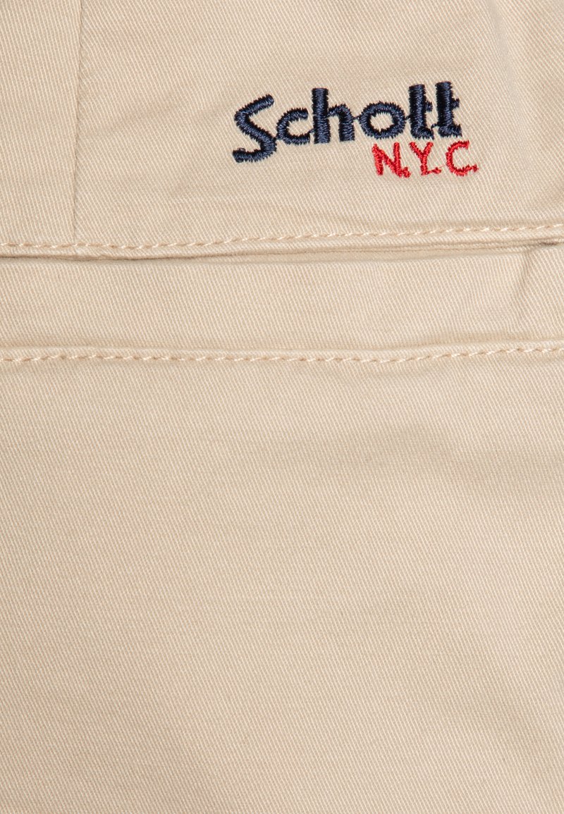 Tissu beige avec une fine texture côtelée. Présente « Schott NYC » brodé en bleu marine et rouge, positionné sur le côté gauche.