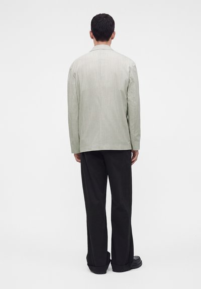 Homme debout de dos portant une veste gris clair, un pantalon noir à jambes larges et des chaussures noires sur fond blanc.