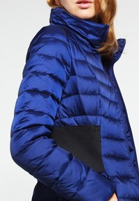 Blauwe pufferjack met hoge kraag, ritsluiting en zwarte elastische pocketdetails. Beschikt over doorgestikte secties en een glanzende, gladde textuur.