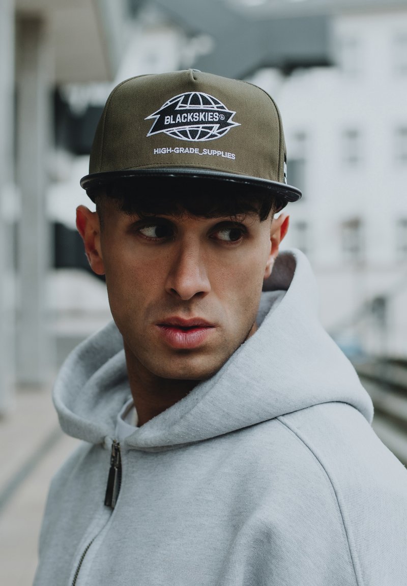 Gorra verde oliva con visera negra, que presenta un logo y texto en relieve en blanco. Lleva puesta una sudadera con cremallera de color gris claro.