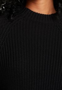 Pull en maille noire avec un motif texturé, encolure côtelée et manches raglan pour une coupe décontractée.