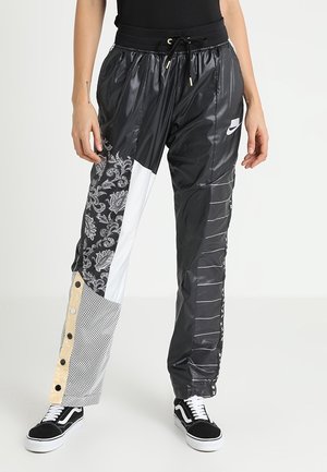 Kobieta nosząca czarne patchworkowe spodnie dresowe Nike z wzorami paisley, siatki i pasków, połączone z czarno-białymi sneakersami.