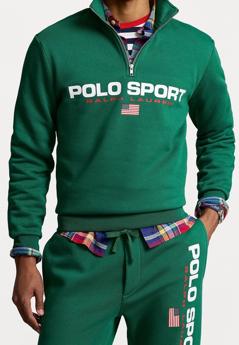 Homme portant un sweat-shirt vert Polo Sport Ralph Lauren et un pantalon de survêtement assorti avec des logos du drapeau américain, superposé à une chemise à carreaux colorée.