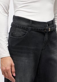 Schwarze Denim-Hose mit einem Gurtbund, Knopfverschluss und seitlichen Taschen. Der Stoff weist eine leichte Ausbleichung auf, um Dimension und Textur zu geben.