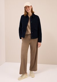 Kvinna som ler, iförd beige mössa, svart knäppt jacka, beige vida byxor och beige sneakers, stående mot en enkel beige bakgrund.