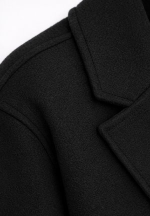 Zbliżenie na ramię i kołnierz czarnego wełnianego płaszcza z widocznymi przeszyciami i fakturowanym materiałem.