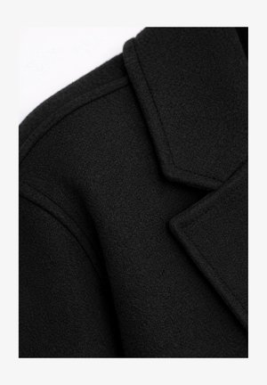 Nahaufnahme von Schulter und Kragen eines schwarzen Wollmantels mit sichtbaren Nähten und strukturiertem Stoff.