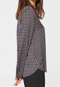 Kvinna iklädd en långärmad, svart blus med ett vitt och rosa rutmönster, tillsammans med svarta byxor, visad från sidan.