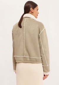 Kurtka z imitacji zamszu w jasnym zielonym kolorze z białymi wykończeniami, z klasycznym kołnierzykiem i widocznymi szwami, o krótkim kroju i miękkiej fakturze.