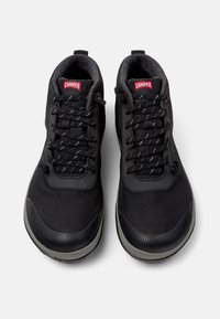 Zapatillas deportivas negras con parte superior de tela texturizada, cordones y una suela de goma gris. Presenta un logo rojo de CAMPER en la lengüeta.