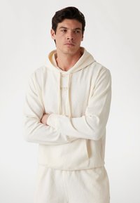 Krémfarvet hætte sweatshirt med et tekstureret bladmønster, med en frontlomme og snørebånd, vist på en model med krydsede arme.