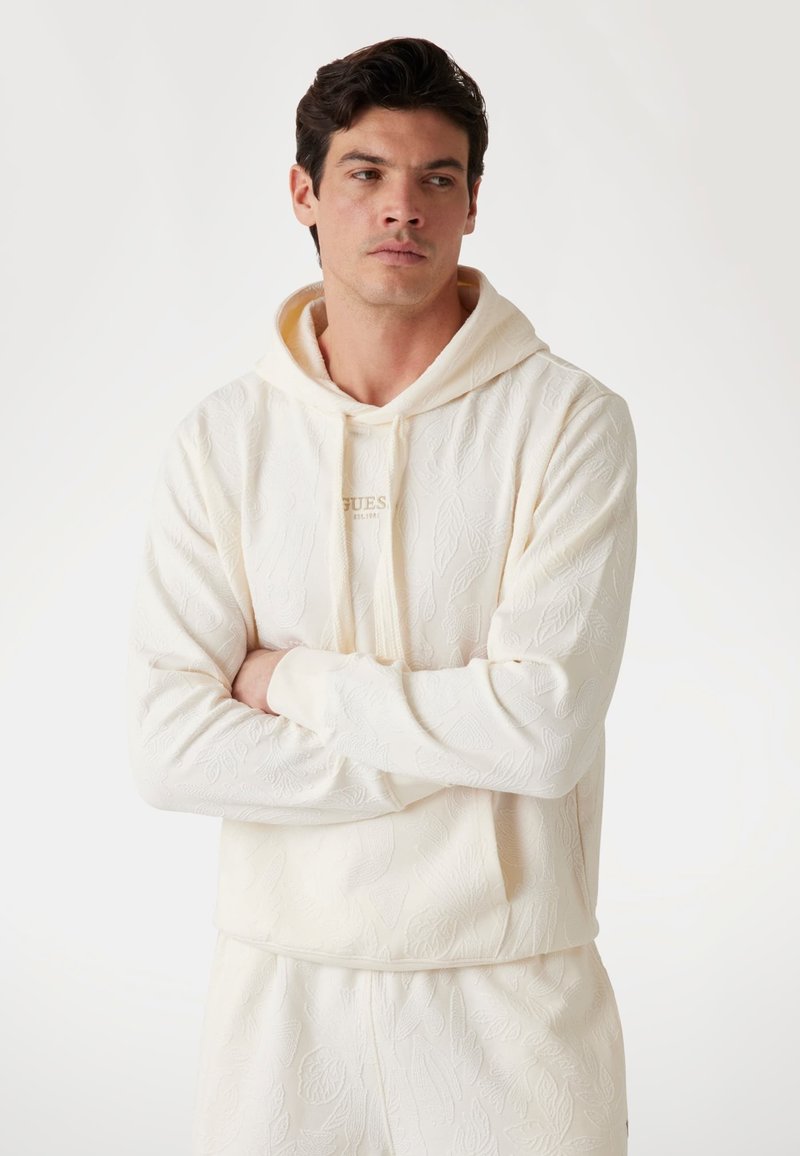 Krémfarvet hætte sweatshirt med et tekstureret bladmønster, med en frontlomme og snørebånd, vist på en model med krydsede arme.