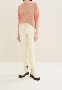 Femme portant un haut corail texturé à manches longues, un pantalon crème à cordon de serrage et des bottines crème à semelles noires, debout devant un fond uni.