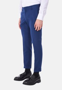 Pantaloni sartoriali blu con vestibilità slim, realizzati in tessuto liscio. Presentano delle pieghe frontali e orli arrotolati. Abbinati a mocassini neri e calzini.
