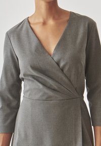Grå omslagsklänning i texturerad tyg med djup V-ringning och trekvartslånga ärmar, med en rynkad midjedetalj.