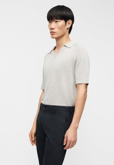 Homme aux cheveux courts noirs portant un polo à manches courtes gris clair et un pantalon noir, debout devant un fond blanc uni.