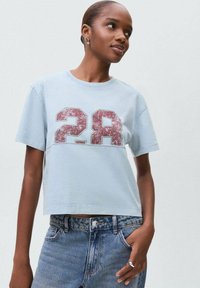 Mladá žena v světle modrém zkráceném tričku s opotřebovaným červeným potiskem "28", oblečená v modrých džínech s vysokým pasem, s rukou v kapse, dívající se doprava.