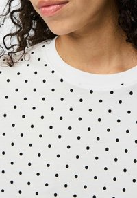 Camisa blanca de manga corta con cuello redondo, que presenta un patrón de lunares negros espaciados de manera uniforme en la parte frontal, hecha de una tela suave.