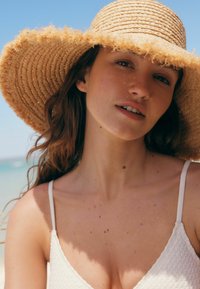 Sombrero de paja de ala ancha con borde deshilachado, llevado sobre una camiseta sin mangas de color claro y textura. El fondo muestra una escena de playa difuminada.
