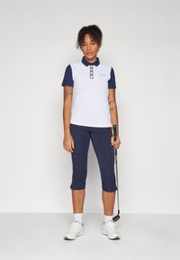 Λευκή και ναυτική κοντομάνικη μπλούζα πόλο με κουμπιά, σε συνδυασμό με ναυτικά κοντά παντελόνια. Το μοντέλο κρατάει ένα γκολφ, ντυμένο με λευκά αθλητικά παπούτσια.