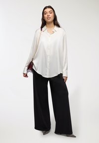 Camicia bianca con bottoni, maniche lunghe, vestibilità comoda, abbinata a pantaloni neri a gamba larga e scarpe decorate. Design semplice e pulito.