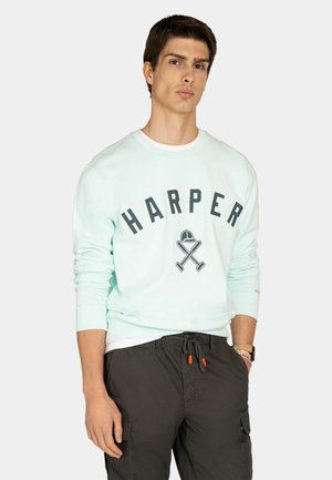 Joven con sudadera celeste con el texto "HARPER" y gráficos de bates de béisbol cruzados y gorra, combinada con pantalones cargo negros.