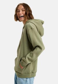 Olivgrön hoodie med justerbar dragkedja, ficka framtill och ribbade mudd. Materialet verkar mjukt med en avslappnad passform. Buren med ljusblå jeans.