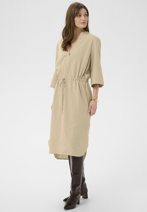 Naine, kes kannab beežikat midi-kleit kolmeveerandvarrukatega, vööga rõhutatud talje ja tumedate põlveni ulatuvate saapadega, seisab tavalise tausta ees.