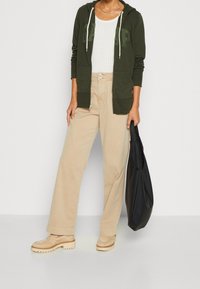 Sudadera verde oliva con cremallera sobre una blusa blanca de canalé, combinada con pantalones de pierna ancha beige y zapatos gruesos beige, sosteniendo un bolso tote negro.
