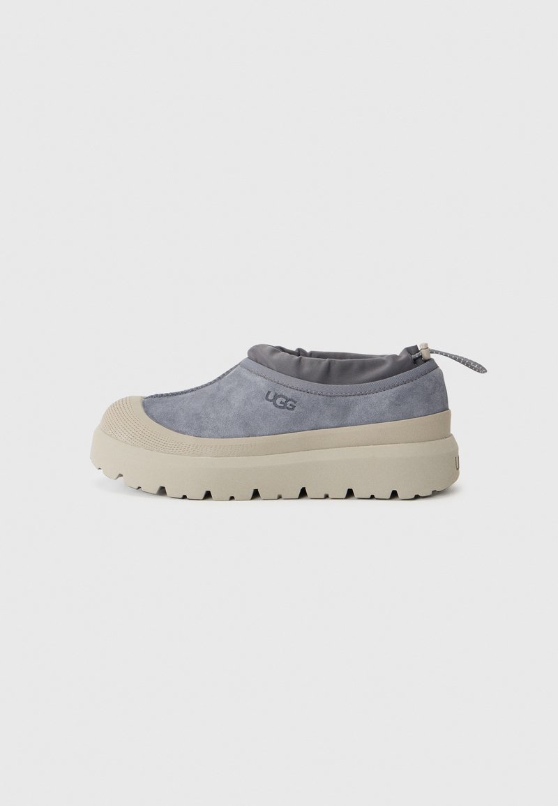 Sivé semišové slip-on topánky so svetlo béžovou gumovou podrážkou, zaoblenou špičkou a elastickým golierom na členku s poutkom pre jednoduché obúvanie.