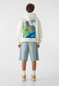 Krēma krāsas kapučjakas ar lielu, krāsainu Amalfi krasta grafiku uz muguras, kopā ar gaiši ziliem džinsa šortiem un bēšām sporta kurpēm.