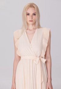 Robe cache-cœur pêche pâle avec des manches à volants, une taille à nouer et une jupe plissée. Tissu doux et léger avec une texture lisse.