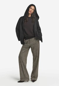 Svart huva cardigan över en mörk tröja, parat med mönstrade vida byxor i brunt och beige. Långt, vågigt hår och klackskor.