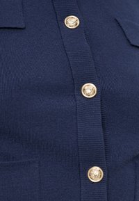 Gros plan sur un cardigan en maille bleu marine avec des boutons décoratifs dorés et une petite poche sur le côté gauche.