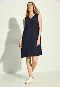 Vestido sin mangas de color marino con escote en V, con un ligero fruncido en los hombros y un corte relajado, combinado con zapatillas blancas. Textura de tela suave.