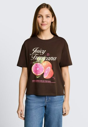 Młoda kobieta nosząca ciemnobrązowy T-shirt z grafiką grejpfruta i napisem „Juicy Tropicana”, w zestawieniu z niebieskimi dżinsami, stojąca na białym tle.